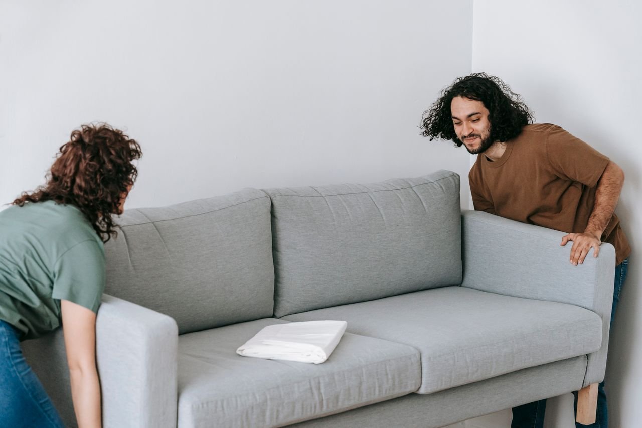 Content ethnic man with anonymous woman carrying comfortable gray couch in living room near white wall during furniture rearrangement at home