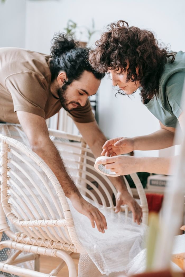A couple collaborates to wrap a chair with bubble wrap for protection during a move.